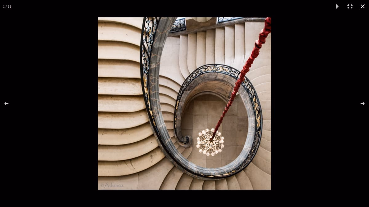 Vue plongeante sur un escalier de pierre en colimaçon, dont la rampe en fer forgé dessine une spirale autour d'un lustre.