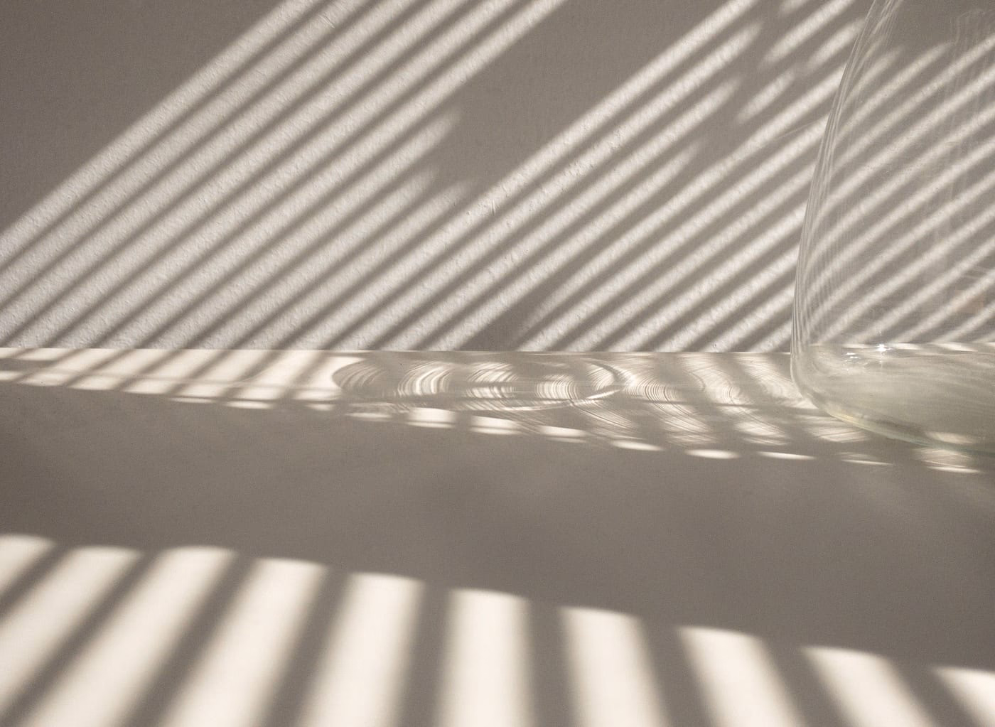 Ombres de lamelles de store projetées en diagonale sur un mur clair, à côté d'un vase en verre.