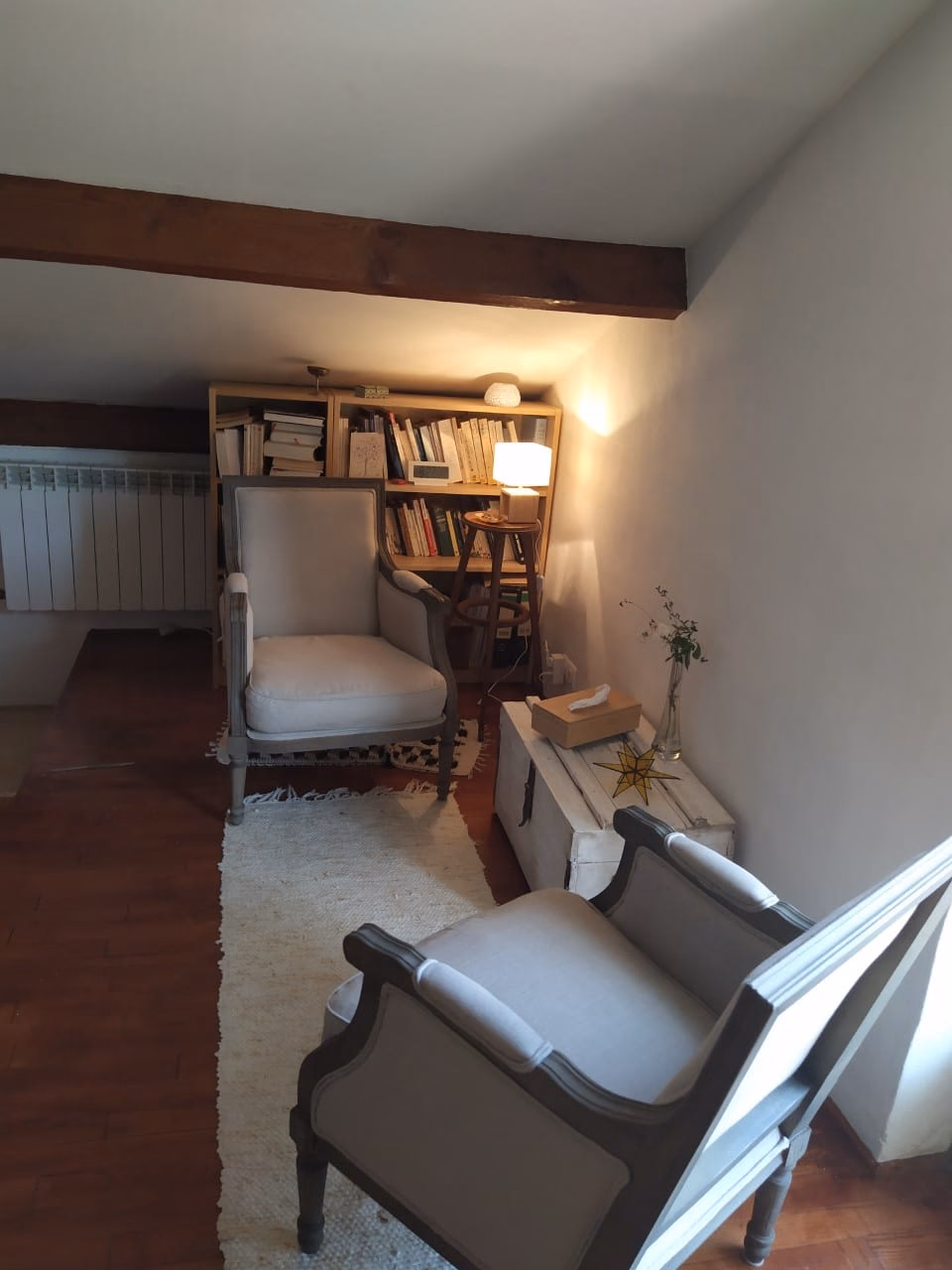 Deux fauteuils gris clair se faisant face sur un tapis de laine, devant une bibliothèque et une lampe allumée, sous des poutres apparentes.