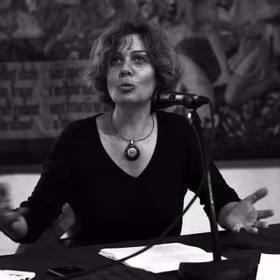 Joëlle Beurier en noir et blanc, assise devant un micro, les mains ouvertes, en train de s'adresser à un auditoire.